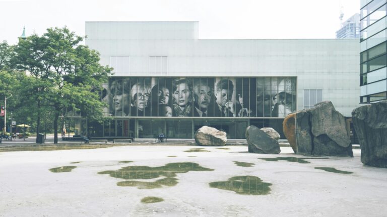 Outdoor scene of a modern museum facade with rocks and large portraits on display.