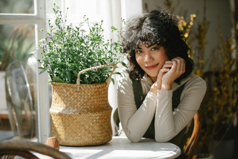 Young smiling Hispanic female in casual clothes sitting at table with basket of flowers and looking at camera